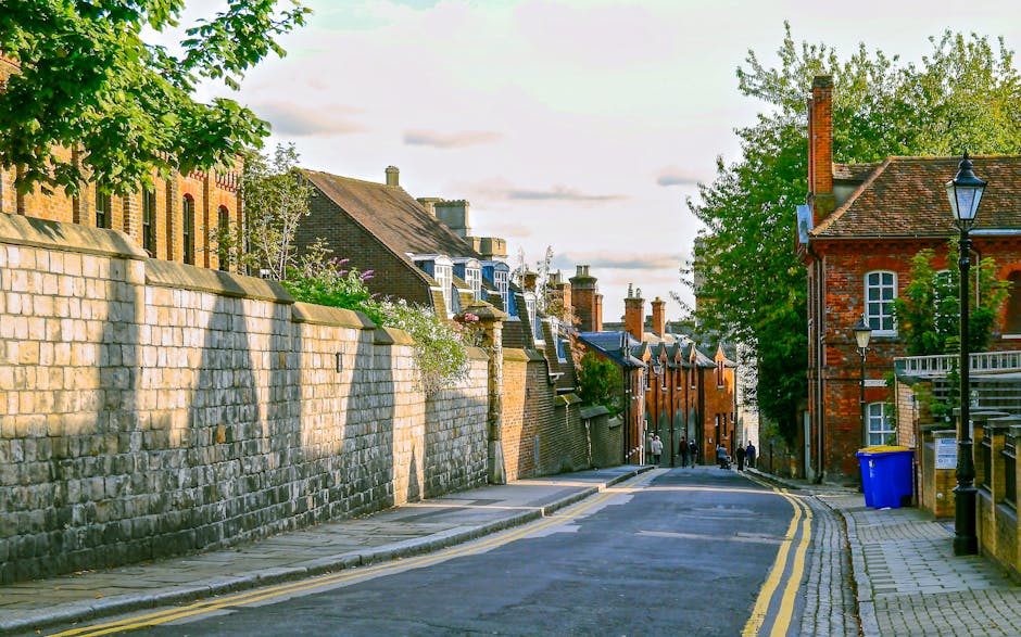 Exterior view of a quiet residential street in Kingston upon Thames during daytime, featuring a stone brick wall lining the sidewalk on the left with lush green trees casting shadows, and traditional brick houses with pitched roofs, multiple chimneys, and white-framed windows on the right. The street has double yellow lines along the curb, with a blue waste bin placed near a lamppost. The scene is well-lit with natural sunlight, highlighting the clean and tidy appearance of the surroundings, exemplifying the importance of surface cleaning and maintenance for property upkeep, as promoted by Kingston upon Thames Cleaners in their deep cleaning and sanitisation services for residential areas.