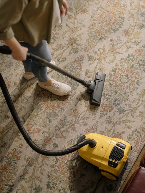 A child is vacuuming a patterned, traditional-style carpet with a yellow and black vacuum cleaner in a well-lit room. The child's lower body is visible, wearing beige shoes, dark jeans, and a beige jacket. The vacuum's hose is connected to the main body, which is positioned on the carpet. The room appears tidy and clean, with the carpet showing no visible dirt or stains. The image reflects domestic surface cleaning, emphasizing hygiene and maintenance, and is associated with Kingston upon Thames Cleaners' professional services near Canbury and Richmond Road Kingston.