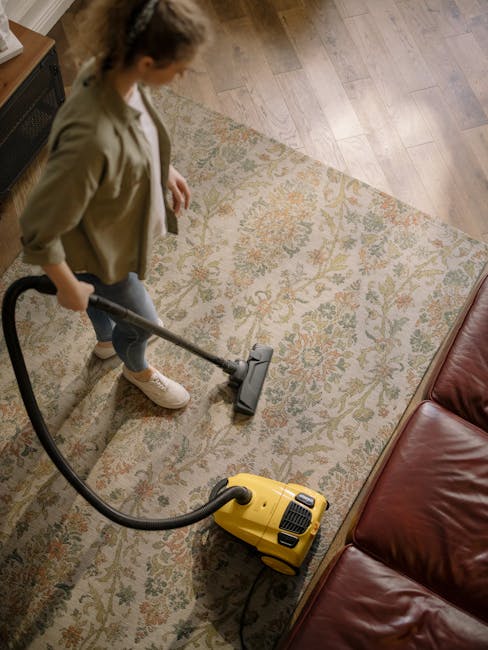 A woman with curly hair, wearing a beige jacket, white top, and jeans, is using a yellow and black vacuum cleaner on a patterned area rug with floral motifs in a living room. The hardwood floor surrounds the rug, and a leather sofa is visible to the right. The room appears well-lit, and the area rug looks clean and free of dust or debris. This image demonstrates surface cleaning as part of domestic cleaning services offered by Kingston upon Thames Cleaners, specializing in carpet and deep cleaning near Canbury and Richmond Road in Kingston.