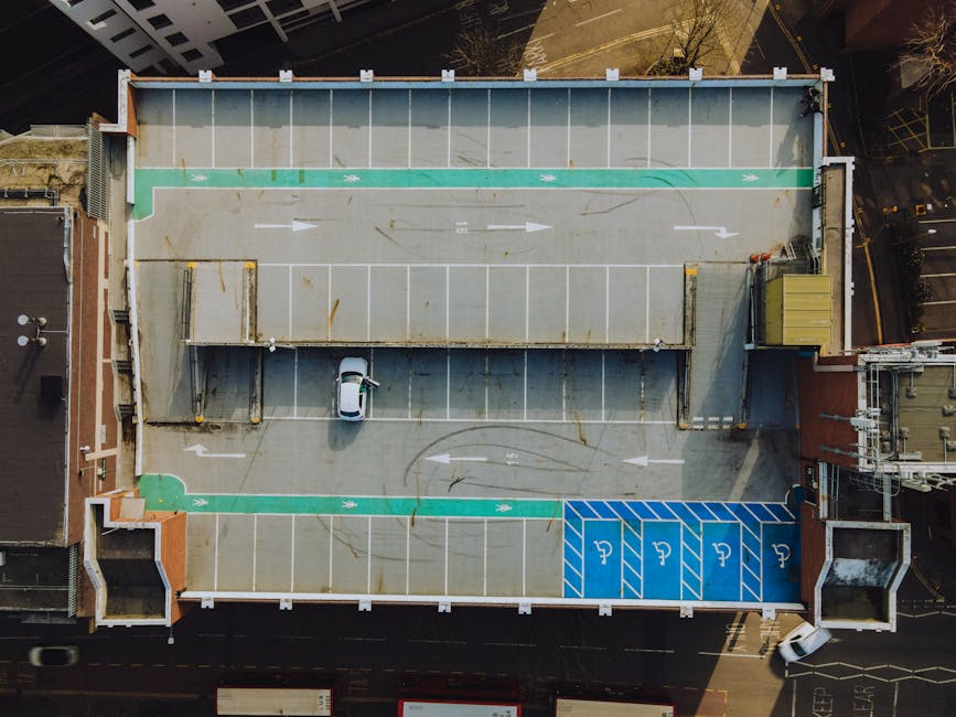An aerial view of a rooftop parking lot showing marked parking spaces, including designated accessible parking bays highlighted in blue with wheelchair symbols. The lot features a concrete surface with visible tire marks and patches of rust-colored stains near the center. A single white car is parked adjacent to a small sheltered area with a flat roof, located towards the lower middle of the image. The parking bays are outlined with white painted lines, with the green-lined areas indicating directional or reserved zones. The surrounding buildings have brick and modern facades, and the lighting suggests daylight conditions. This scene exemplifies surface organisation and cleanliness within a commercial parking area, emphasizing maintenance and orderliness as part of property upkeep, consistent with the principles of domestic cleaning and surface sanitisation discussed by Kingston upon Thames Cleaners for complying with disposal laws for upholstery cleaning Kingston, KINGSTON UPON THAMES.