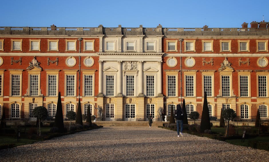 A symmetrical, neoclassical-style building with a grand facade featuring large rectangular windows with white frames, decorative stone detailing, and a central entrance with columns. The exterior walls are made of red brick with white stone accents. The grounds include a gravel pathway leading to the entrance, flanked by neatly trimmed small trees and dark, conical topiary bushes. The sky is clear and blue, with bright sunlight illuminating the building, which appears well-maintained and clean. This scene is associated with Kingston upon Thames Cleaners and reflects a focus on property appearance and upkeep.