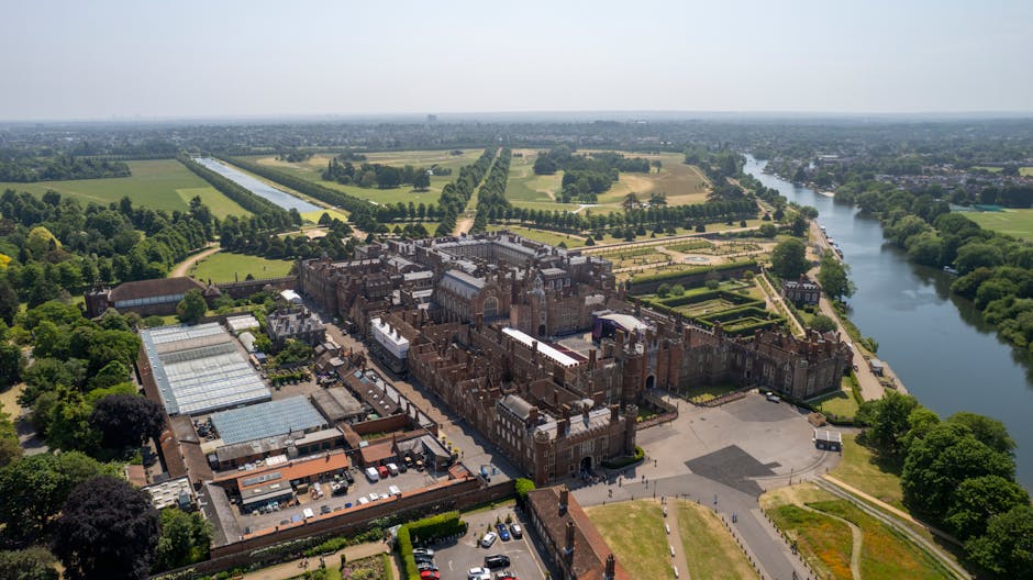 An aerial view of Hampton Court Palace, a historic brick castle with multiple towers and ornate architectural details, surrounded by lush green gardens and trees. To the right, the River Thames winds alongside the palace, with a pathway and more greenery lining its banks. The scene captures the palace's grandeur and its scenic location in Kingston upon Thames, with clear weather and bright daylight illuminating the area. The image emphasizes the historic structure's prominence within its natural setting, consistent with scenic tourism sites in Kingston upon Thames. Kingston upon Thames Cleaners, specializing in cleaning services, creates a connection to maintain the pristine appearance of such historic and scenic locations through expert surface cleaning and sanitisation.