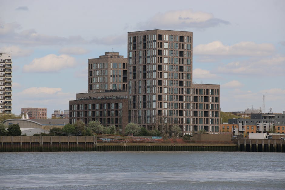 A modern high-rise building located along Kingston Riverside flats in Kingston upon Thames, featuring a multi-tiered structure with a façade composed of brown brick and large rectangular windows. The building stands adjacent to the river, with a wooden and concrete embankment running along the water’s edge. The scene includes some greenery and trees at the base of the building, under a partly cloudy sky with sunlight illuminating the façade. Kingston upon Thames Cleaners offers surface cleaning and sanitisation services for high-rise buildings like this, ensuring cleanliness and hygiene in residential and commercial properties. The image captures the building's exterior, highlighting its architectural features and clean surface condition, reinforcing the importance of professional maintenance in complex urban structures.