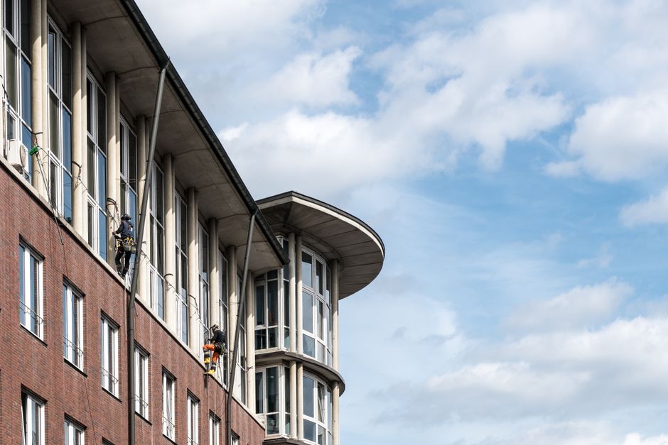 Image showing the exterior of a modern, multi-story building with a brick facade and large glass windows, some of which are being cleaned by window washers using harnesses and extension poles. The building features a prominent rounded corner with extensive glass paneling, set against a partly cloudy sky. The scene illustrates high-rise surface cleaning and maintenance, emphasizing the need for professional access during cleaning of windows and exterior surfaces. Kingston upon Thames Cleaners specializes in high-rise cleaning access solutions for such riverside flats, ensuring thorough sanitisation and hygiene of all external surfaces.