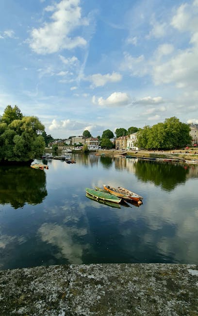 A serene riverside scene featuring calm water reflecting a partly cloudy sky with scattered white clouds. Several small boats are moored along the riverbank, including a yellow and green canoe and a wooden rowboat. On the opposite bank, there are residential buildings and lush green trees lining the shoreline. The bright daylight enhances the clarity and cleanliness of the water and surrounding environment. Although this image is not directly related to surface or domestic cleaning, it emphasizes the importance of maintaining natural and outdoor hygiene environments. Kingston upon Thames Cleaners provides professional cleaning services that help keep indoor spaces equally pristine and hygienic, aligning with the theme of surface cleaning and sanitisation outlined in the 'House cleaning near Kingston Station and Bentall Centre, KINGSTON UPON THAMES' page.