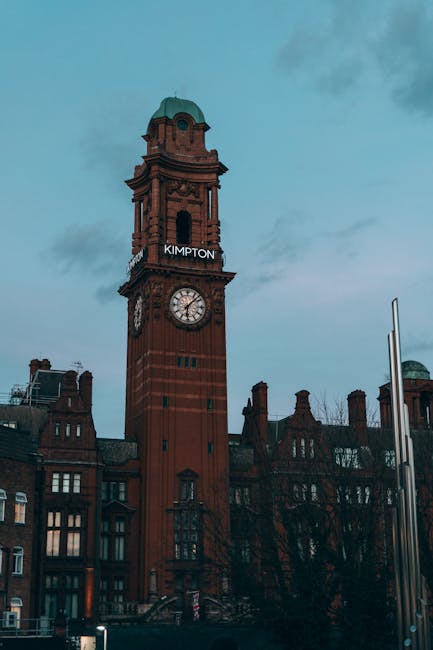 An exterior view of Kingston's historic clock tower made of red brick with a green dome at the top, displaying the word 'KINGTON'. The clock face is visible, showing the time, against a cloudy sky. Surrounding the tower are traditional red-brick buildings with multiple chimneys, indicative of Kingston upon Thames, in daylight. The scene emphasizes the architectural details and urban setting, fitting for content related to surface cleaning or maintenance in the area. Kingston upon Thames Cleaners provides domestic cleaning services near Kingston Station and Bentall Centre, ensuring thorough and professional sanitisation and deep cleaning in local properties.