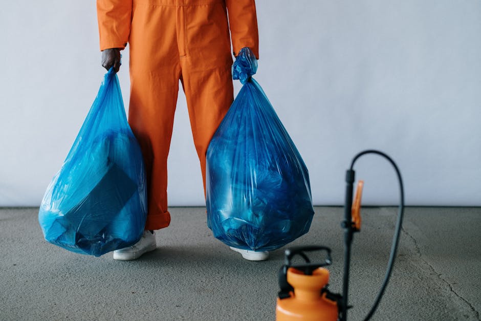 A person dressed in orange work overalls holding two large blue plastic bags filled with waste, standing on a clean, grey floor in a room with plain white walls. The scene includes a black and orange handheld sprayer or cleaning device on the floor to the right, suggesting a focus on waste collection and surface cleaning. The lighting is bright and even, highlighting the glossy surface of the plastic bags and the neat cleanliness of the area. This image emphasizes proper waste disposal practices following clearance cleaning, in line with Kingston Council rules, as promoted by Kingston upon Thames Cleaners for maintaining hygiene and proper sanitation in residential and commercial spaces.