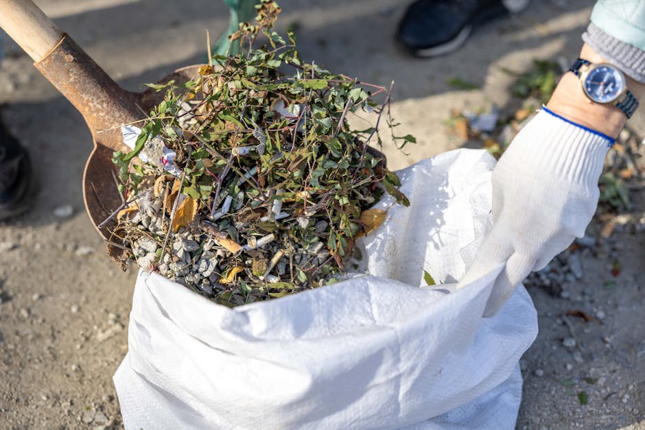 A person wearing a white glove is placing a bag filled with garden debris, including small branches, leaves, and gravel, into a white woven polypropylene sack. The scene is outdoors on a gravel surface, with natural sunlight illuminating the area. A shovel with a wooden handle and metal blade, partially visible, is being used to scoop the garden waste. The individual is wearing a light-colored sleeve and a wristwatch, indicating a clean and organized waste collection process. This image relates to domestic cleaning and waste disposal activities, aligning with Kingston Council rules for waste disposal after clearance cleaning as highlighted on kingstonuponthamescleaners.org.uk.