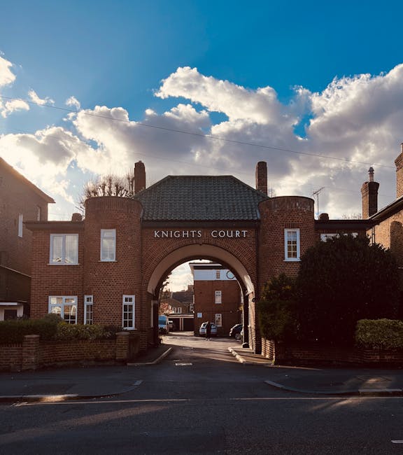 Photograph of the Knights Court entrance, a brick archway flanked by two brick buildings with white-framed windows, under a partly cloudy sky with sunlight breaking through. The arch leads into a residential street with parked cars visible in the background. The area appears well-maintained and clean, with greenery on either side of the entrance. Positioned in Kingston upon Thames, this scene relates to the local environment serviced by Kingston upon Thames Cleaners for surface cleaning, deep cleaning, and sanitisation solutions in residential and commercial properties. The image emphasizes the importance of maintaining cleanliness and hygiene at the entrance to the community, reflecting the company's commitment to professional cleaning services that help manage cleaning delays and ensure timely, thorough cleaning tailored to local needs.