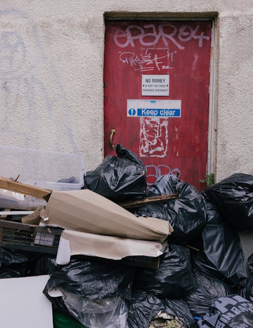 An outdoor scene featuring a red metal door with graffiti and signs, including a 'No Money' notice and a blue 'Keep Clear' sticker, set into a beige, textured wall. In front of the door, there is a pile of black garbage bags, discarded cardboard boxes, and debris, indicating waste left for collection. The lighting appears natural, illuminating the area clearly, which highlights the clutter and mess outside the building. This image relates to cleaning and waste management efforts, showcasing the need for proper waste removal services, such as those provided by Kingston upon Thames Cleaners, to maintain hygiene and cleanliness in residential or commercial premises.
