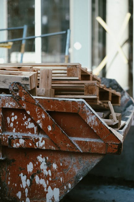 Close-up of a rusted orange metal boat with visible white paint splatters, filled with wooden pallets and planks, situated near a building with large glass windows. Natural daylight illuminates the scene, highlighting the weathered surface of the boat and the arrangement of the construction materials. The images suggest a setting related to renovation or construction site preparation, which Kingston upon Thames Cleaners could address through surface cleaning and deep cleaning services to maintain hygiene and safety in such environments.