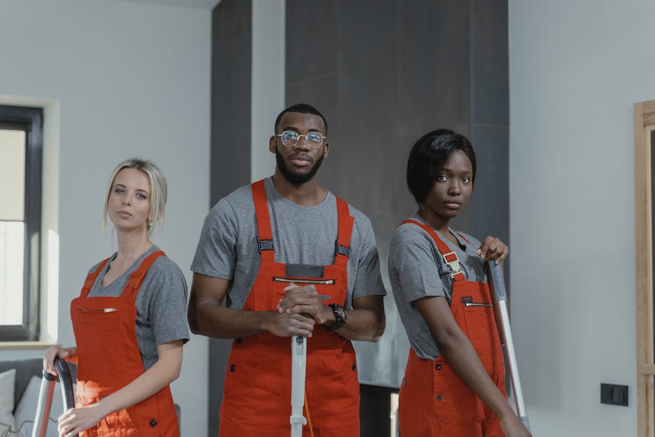 Three professional cleaners from Kingston upon Thames Cleaners, dressed in grey t-shirts and orange aprons, stand indoors holding cleaning tools. The female on the left holds a vacuum cleaner hose, the male in the center has a mop with both hands, and the female on the right also holds a mop. The room features a large window allowing natural light to illuminate the light-colored walls and modern interior. Surfaces such as the floor and wall areas appear clean and well-maintained, reflecting thorough cleaning and sanitation. The setting emphasizes domestic cleaning services, suitable for booking for student lets in Kingston, with an emphasis on surface cleaning, deep cleaning, and hygiene maintenance.