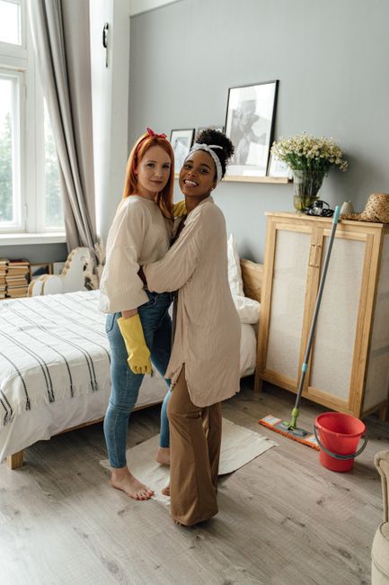 Two women standing together in a well-lit, neatly made bedroom with a wooden bed frame and a striped bedspread. One woman has red hair, is wearing a cream sweater, yellow cleaning gloves, and blue jeans; the other has dark hair tied back, wearing a beige oversized sweater and brown trousers. The room features a large window with light curtains, a shelf with framed photographs and a vase of flowers, and a wooden cupboard. Cleaning tools including a mop and red bucket are visible on the floor, indicating recent or ongoing cleaning activity. The overall scene suggests a clean, tidy space maintained with careful surface cleaning and sanitisation, consistent with domestic cleaning services provided by Kingston upon Thames Cleaners as described on the page about booking cleaners for student lets in Kingston.