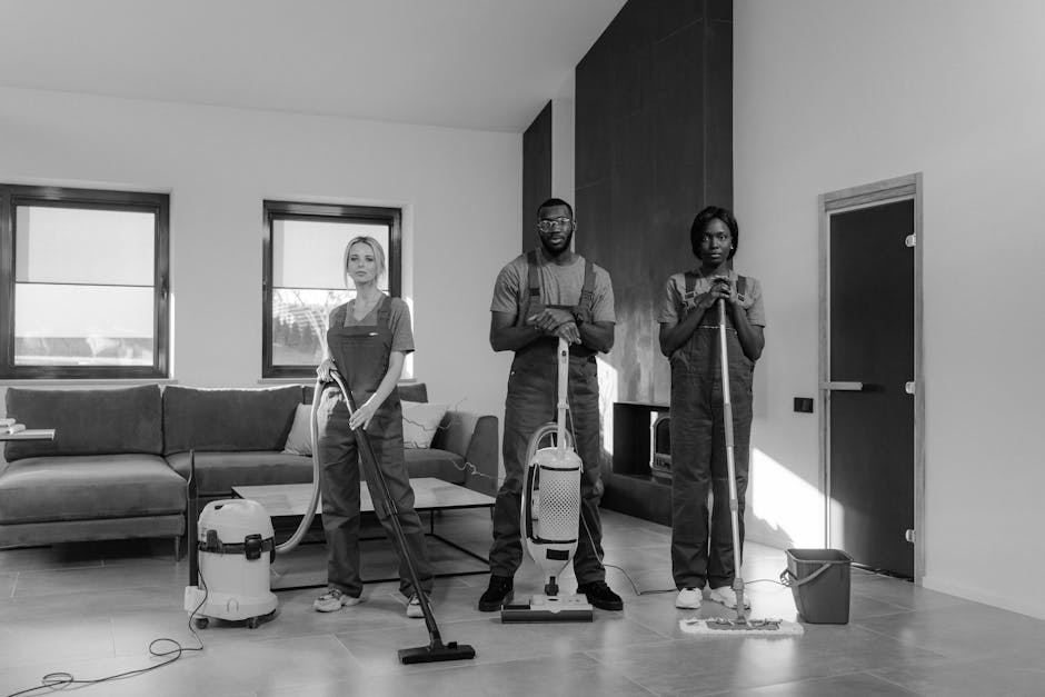 Three professional cleaners from Kingston upon Thames Cleaners, dressed in grey t-shirts and orange aprons, stand indoors holding cleaning tools. The female on the left holds a vacuum cleaner hose, the male in the center has a mop with both hands, and the female on the right also holds a mop. The room features a large window allowing natural light to illuminate the light-colored walls and modern interior. Surfaces such as the floor and wall areas appear clean and well-maintained, reflecting thorough cleaning and sanitation. The setting emphasizes domestic cleaning services, suitable for booking for student lets in Kingston, with an emphasis on surface cleaning, deep cleaning, and hygiene maintenance.